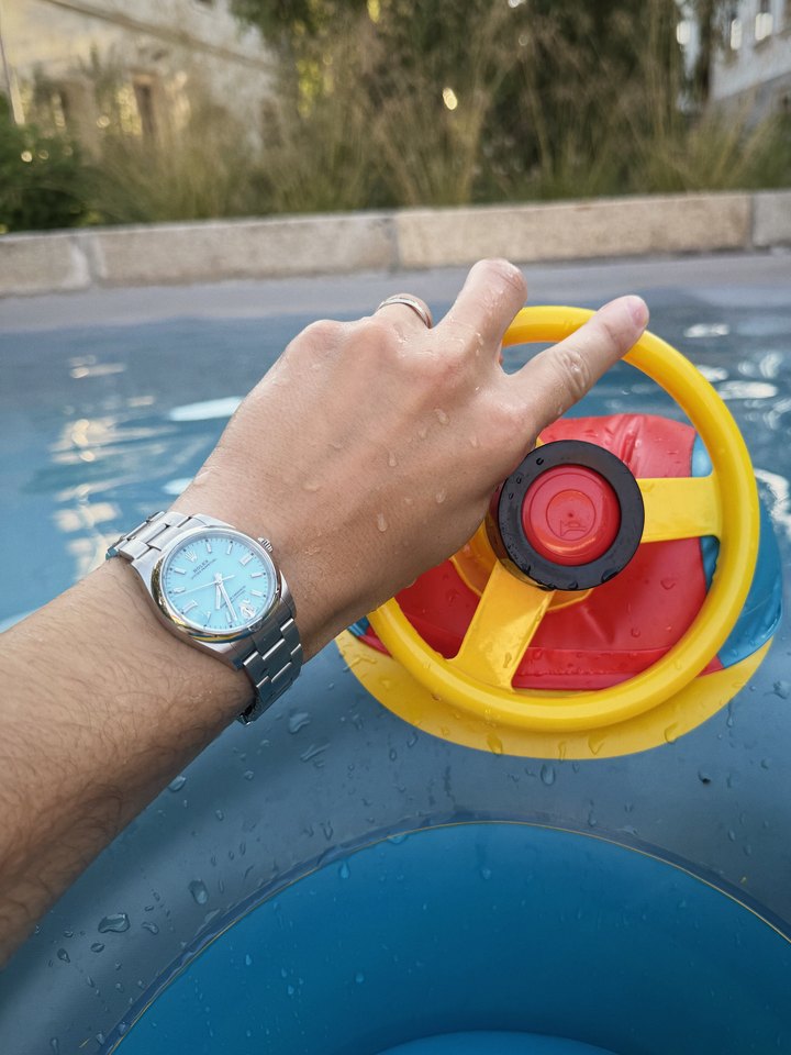 My wrist with a turquoise-dial watch, hand on the yellow steering wheel of a child's pool float.